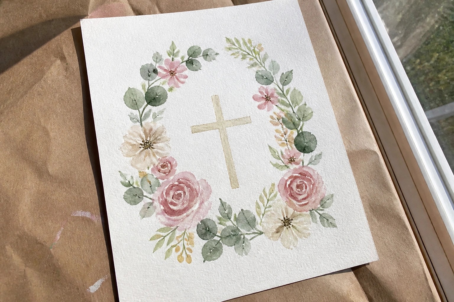 A watercolor painting of pink and white roses with green leaves forming a wreath around a central beige cross on white paper.