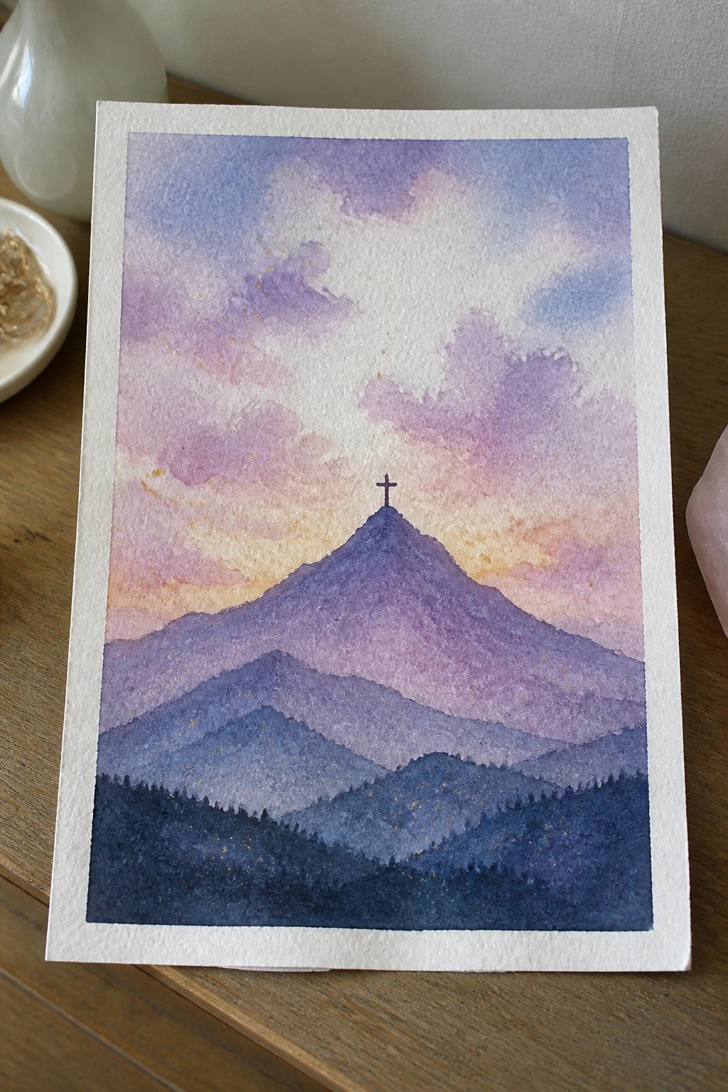 Watercolor painting of layered purple mountains with cross on peak under pastel sky.