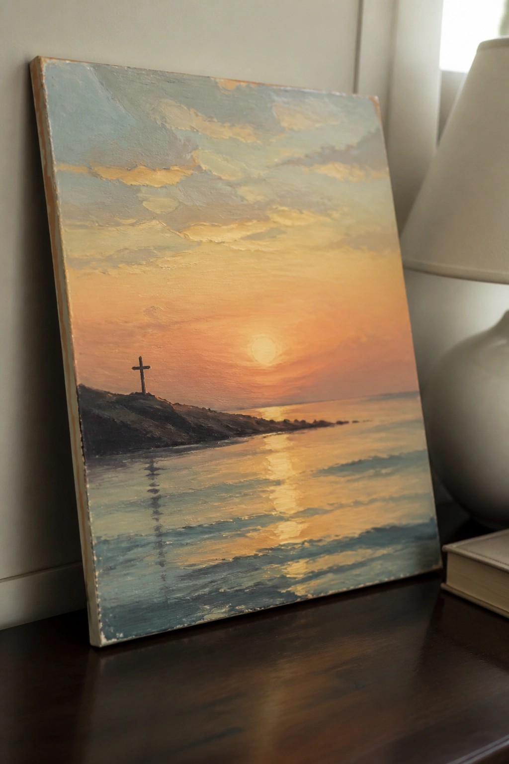 Oil painting of sunset over ocean with cross on hill next to lamp on table.