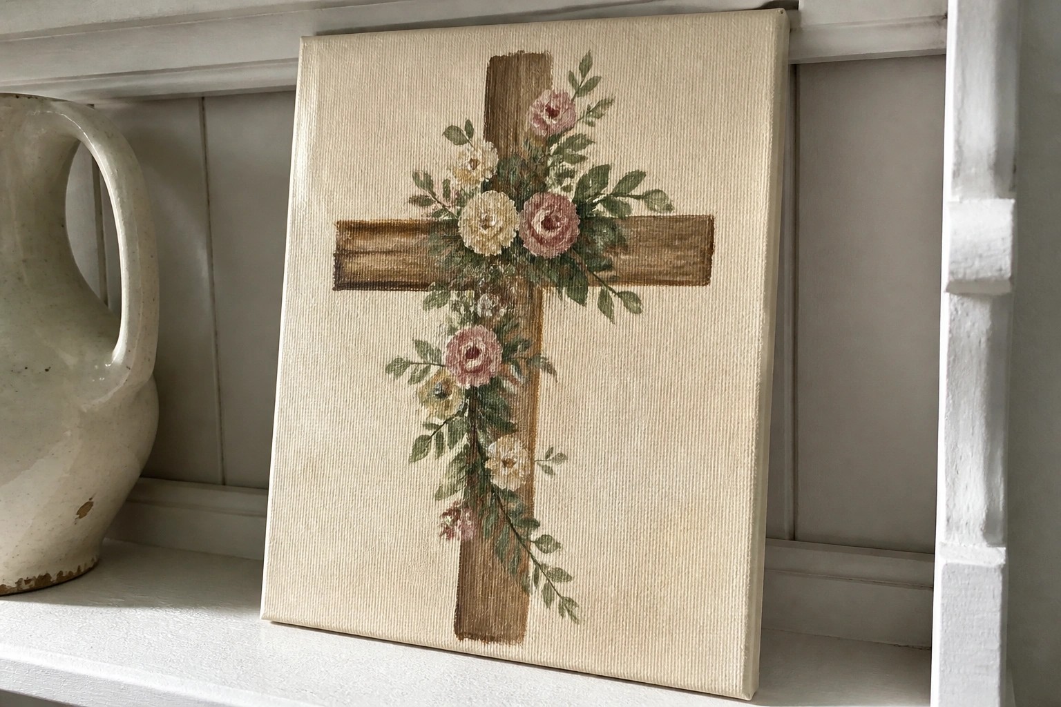 A canvas painting shows a brown wooden cross with pink and white flowers and green leaves arranged across it on a cream background.