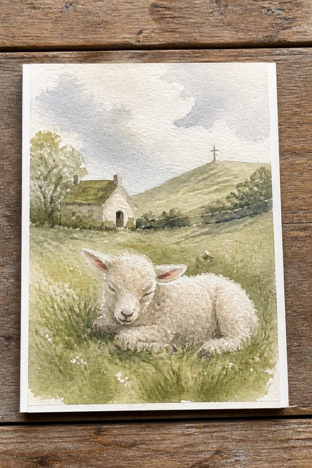 Watercolor of a white lamb resting in grassy field with cottage and hills.