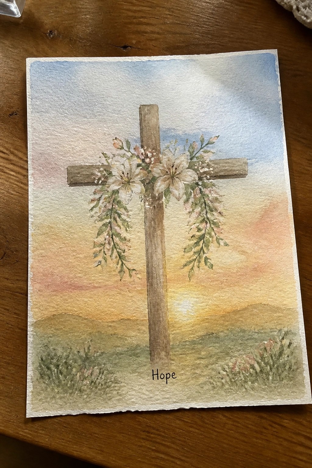 Watercolor of wooden cross with white flowers and vines over sunset landscape labeled Hope.
