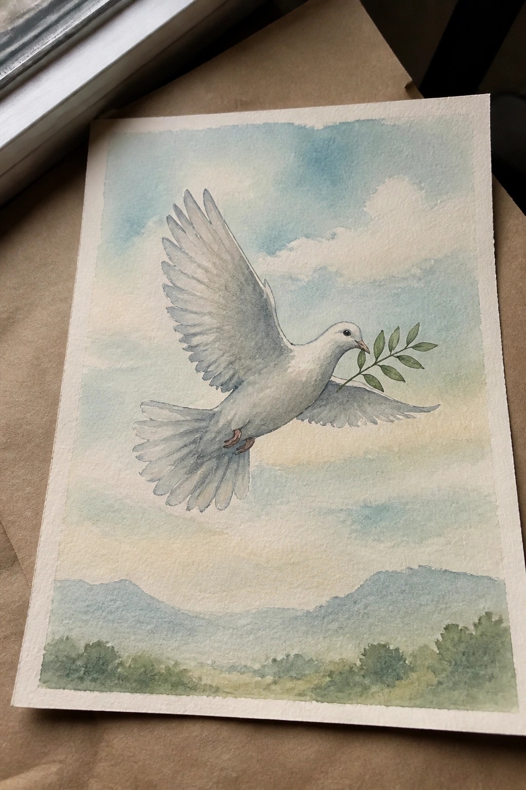 Watercolor of white dove carrying olive branch, flying over misty blue mountains and sky.