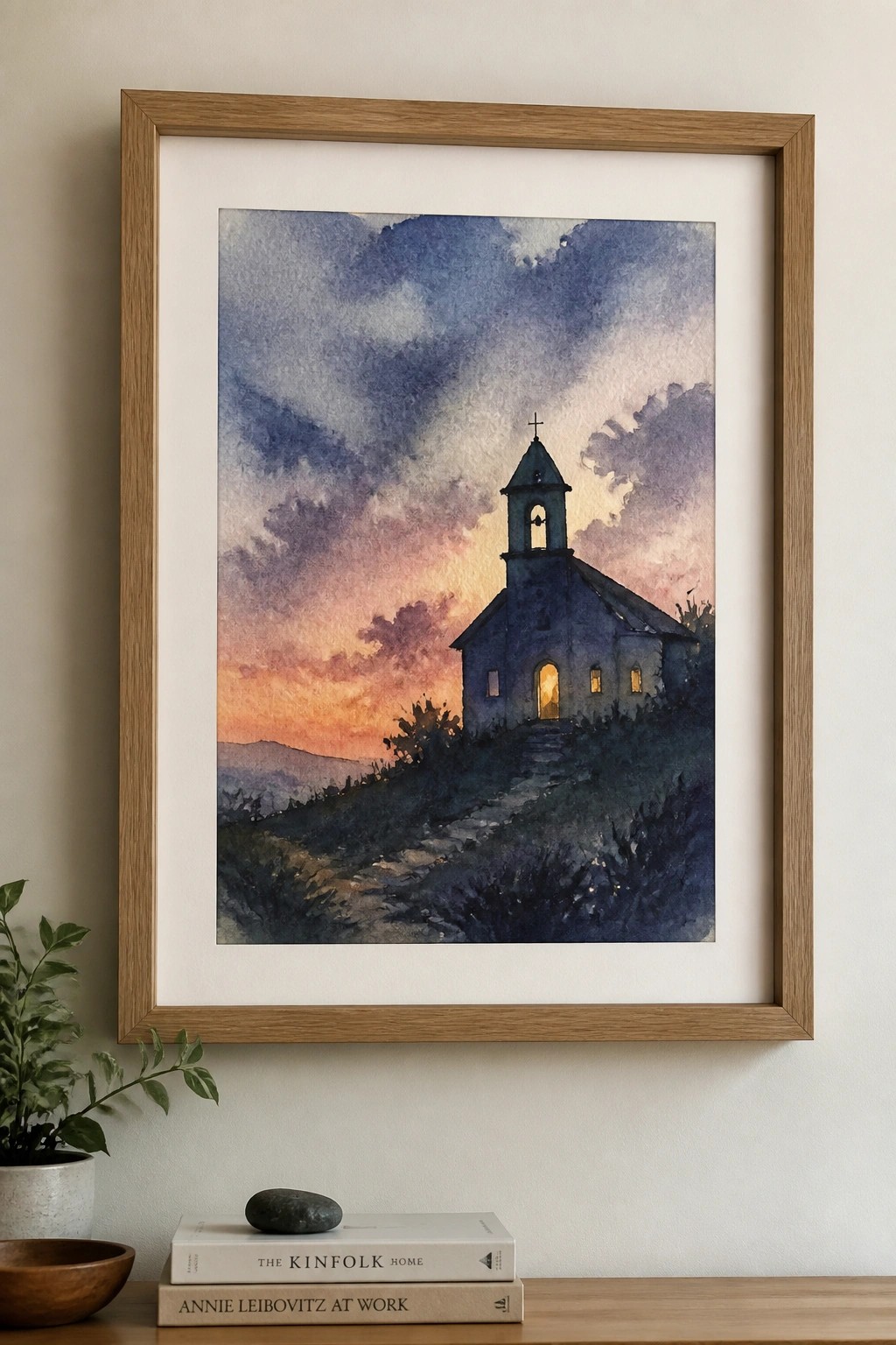 Framed watercolor of church at sunset above stacked books, stone, and potted plant.
