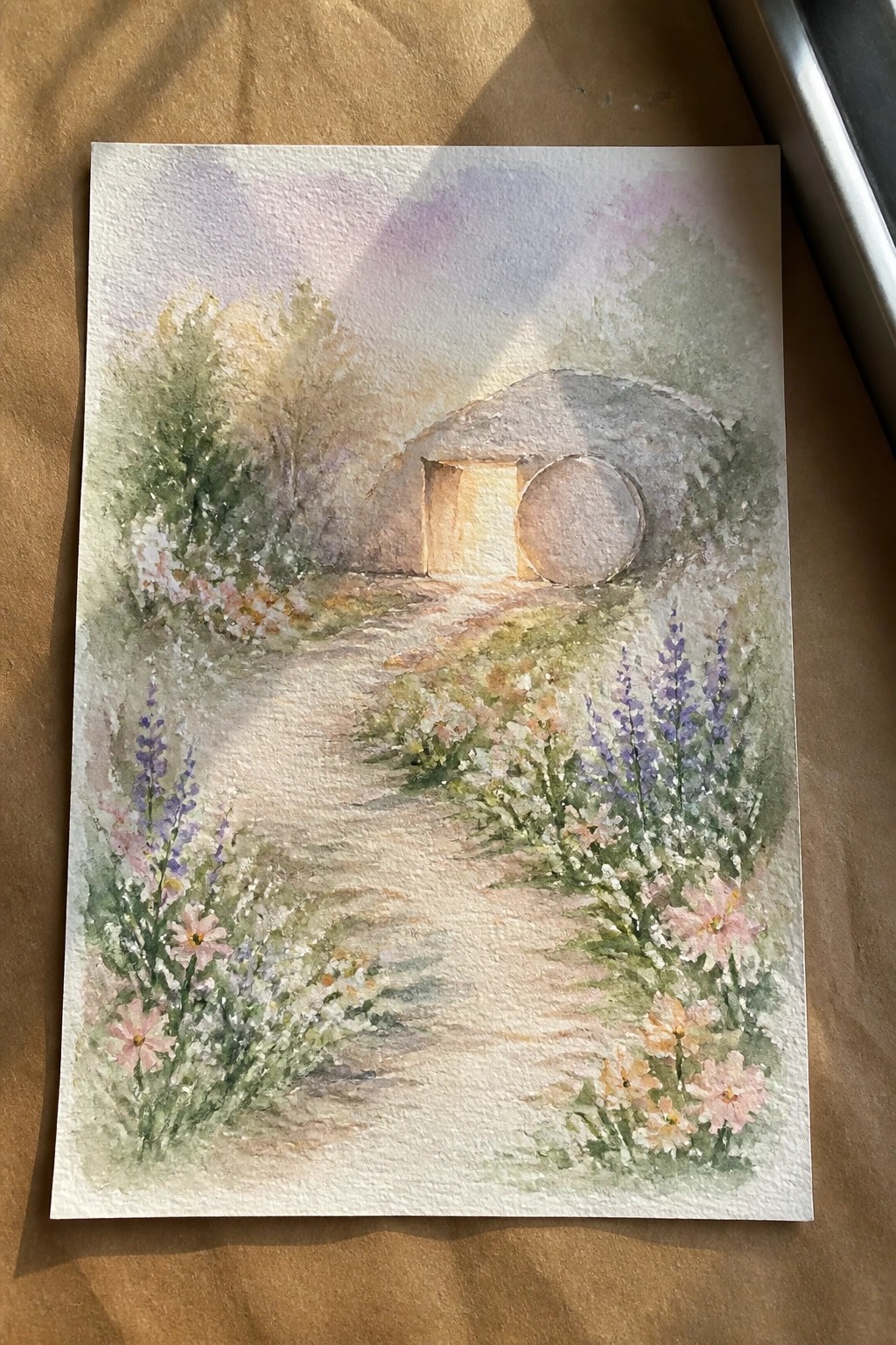 Watercolor painting of flower-lined path leading to glowing stone tomb with open door.