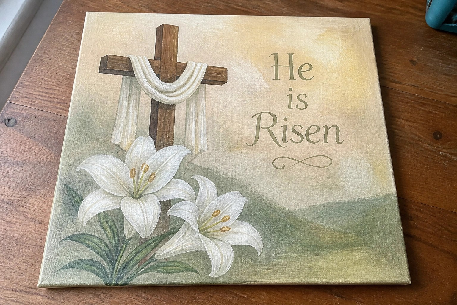 Canvas painting of wooden cross with white cloth, lilies, and "He is Risen" text