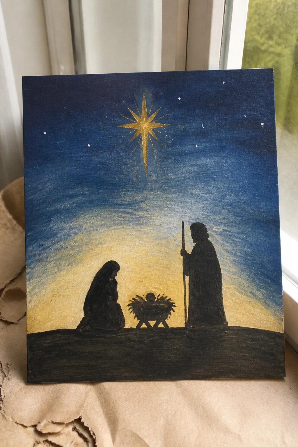 Silhouetted Nativity with kneeling Mary, staff-holding Joseph, manger, and radiant golden star in blue sky.