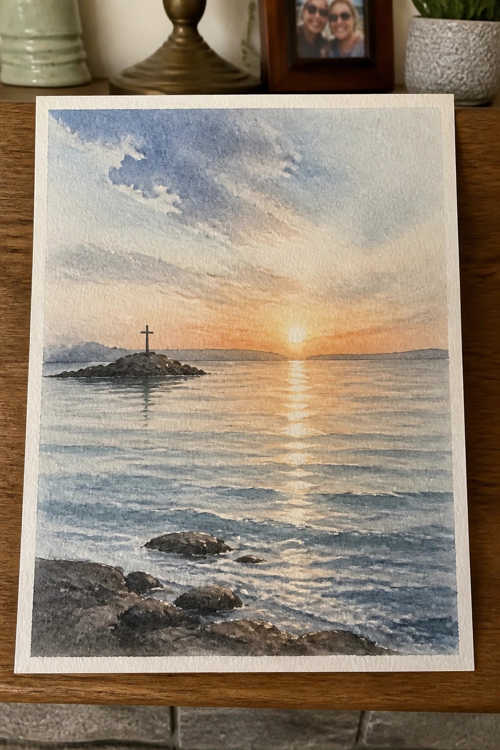 Watercolor seascape of ocean sunset with sun reflection, cross on rocky island, and foreground rocks.