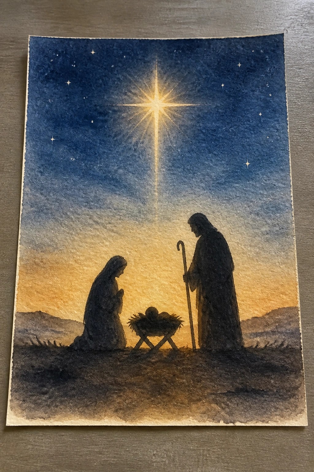 Watercolor nativity with Mary kneeling, Joseph with staff, manger under radiant starlit sky.