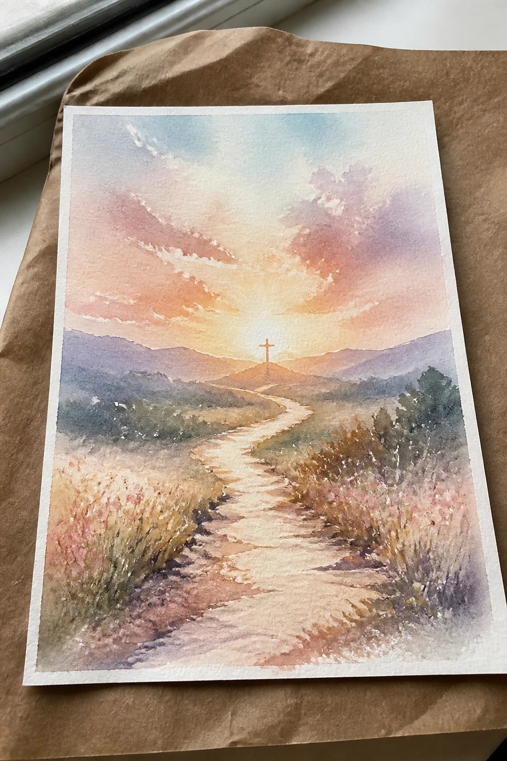A watercolor painting of a winding dirt path through fields leading to a cross on a hill at sunset with mountains behind.