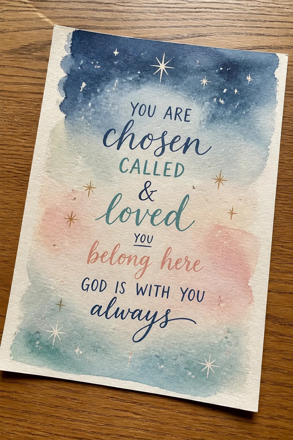 Watercolor card on wood with blue-pink gradient, gold stars, and religious text about being chosen and loved