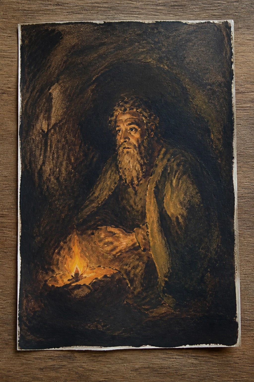 Bearded man in tattered robes sits by a small fire in a dark cave.