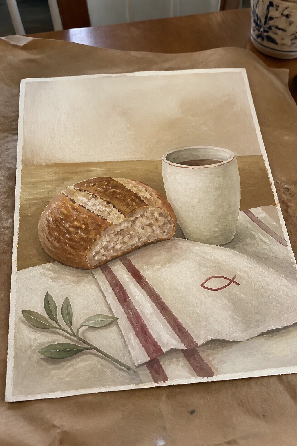 Still life painting of bread loaf, cup, striped cloth with fish symbol and leaves