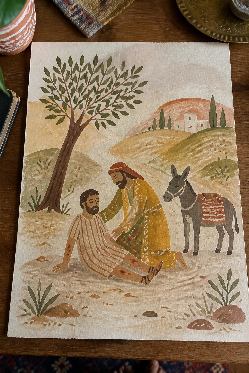 Watercolor shows yellow-robed man aiding injured figure beside donkey under tree in hilly landscape.