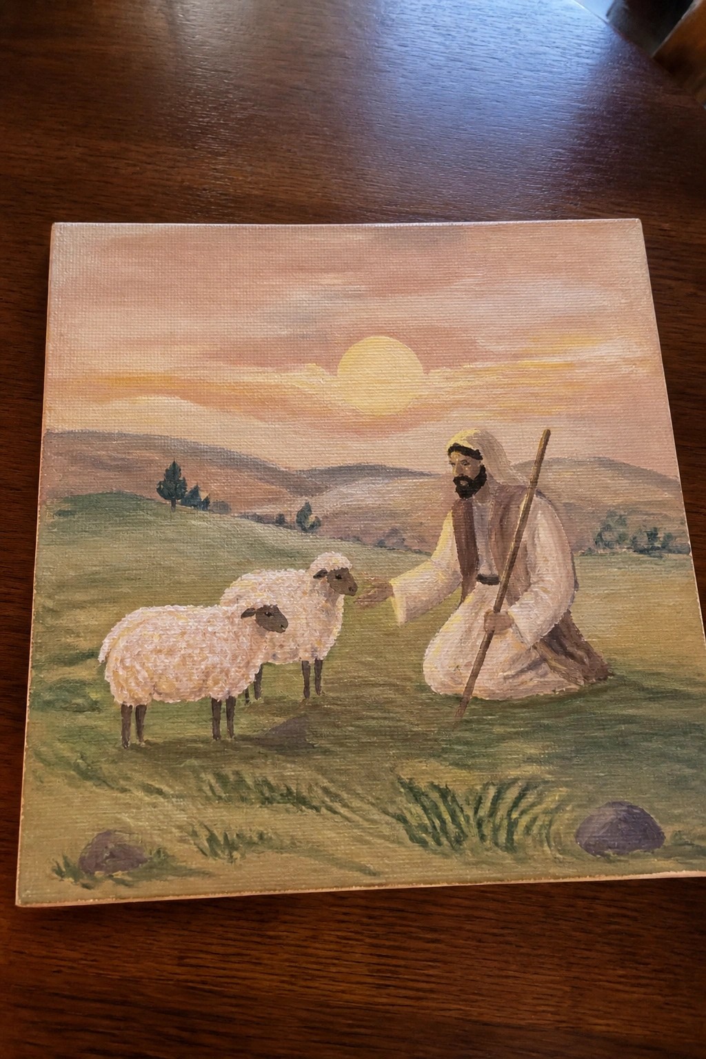 Bearded shepherd in white robes kneels holding staff beside two sheep at sunset.