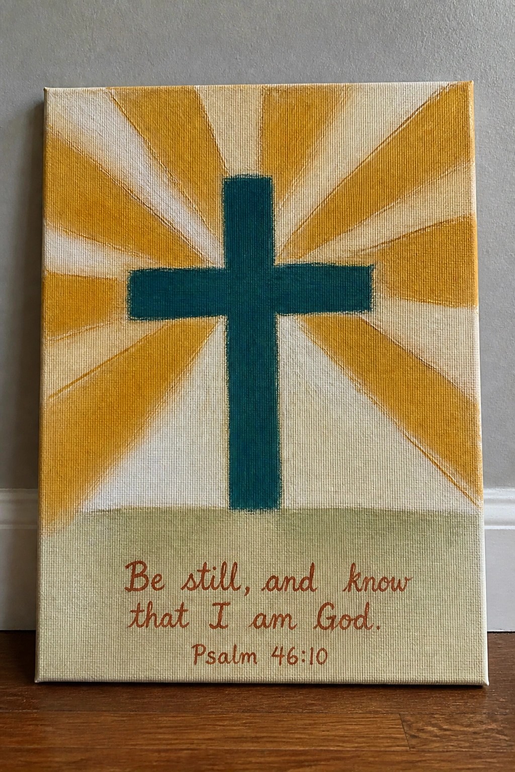 A canvas painting features a dark teal cross centered on yellow and white radiating stripes with a green bottom section containing brown text of Psalm 46:10.