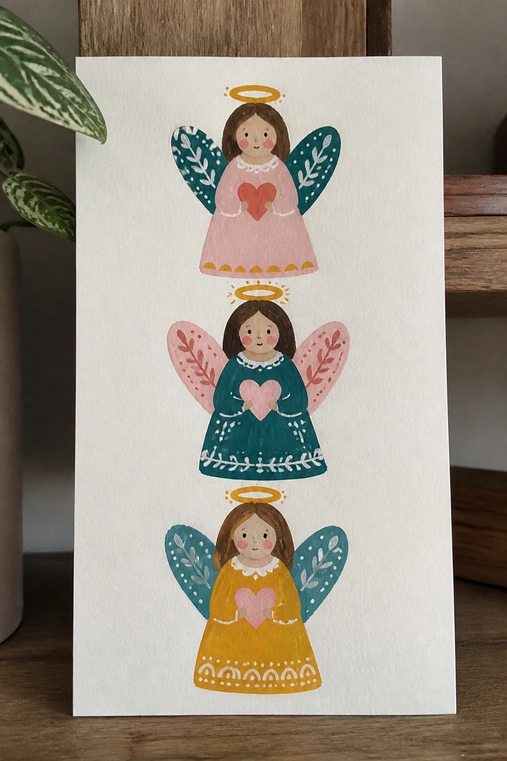 Three stacked angels in pink, teal, and yellow dresses with halos and hearts.