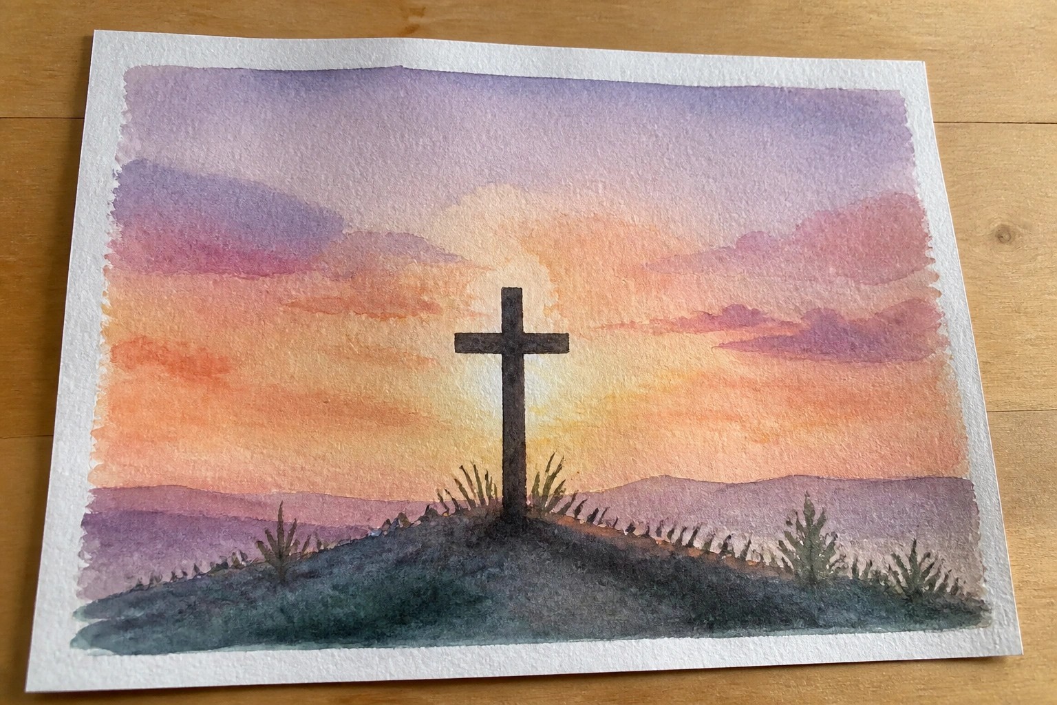 Watercolor painting of a dark cross on hills under a blended orange-purple sunset sky.