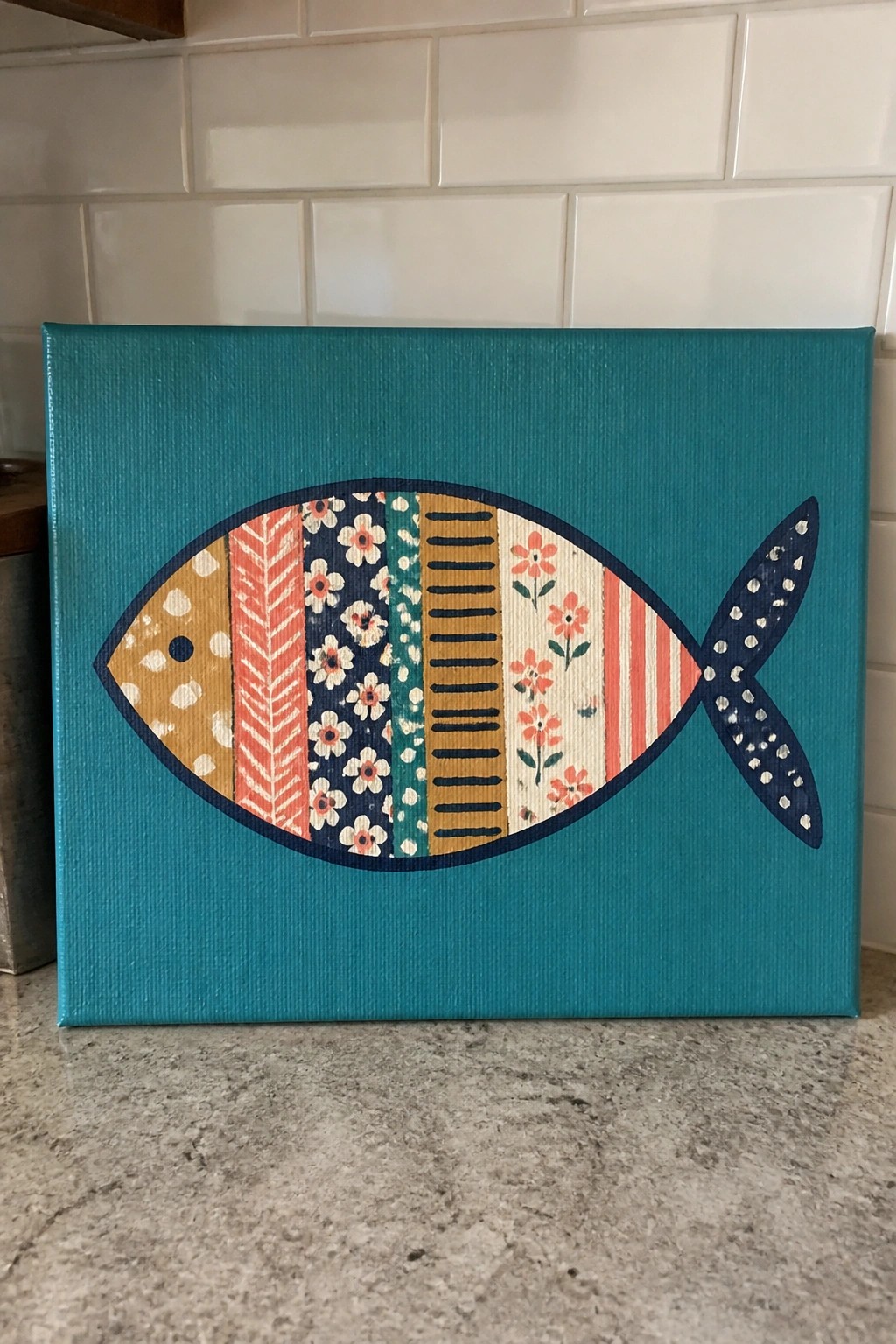 Colorful patterned fish painting on teal canvas against white tile backsplash