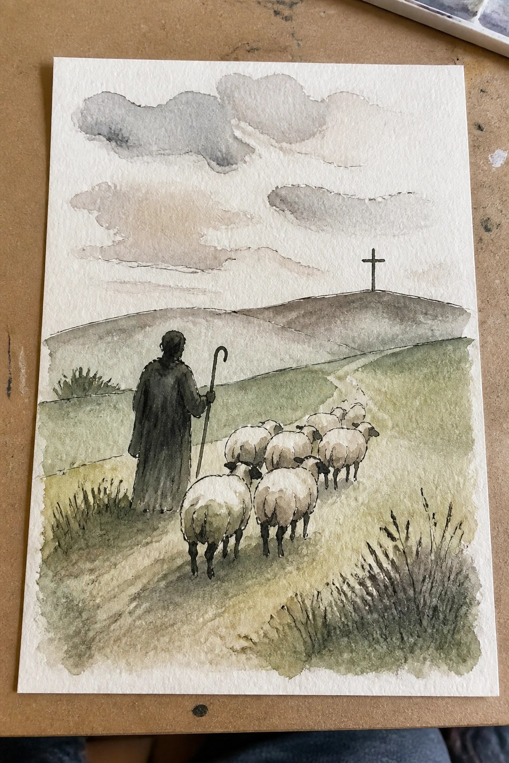 Watercolor of robed shepherd with staff herding sheep along path toward hilltop cross.