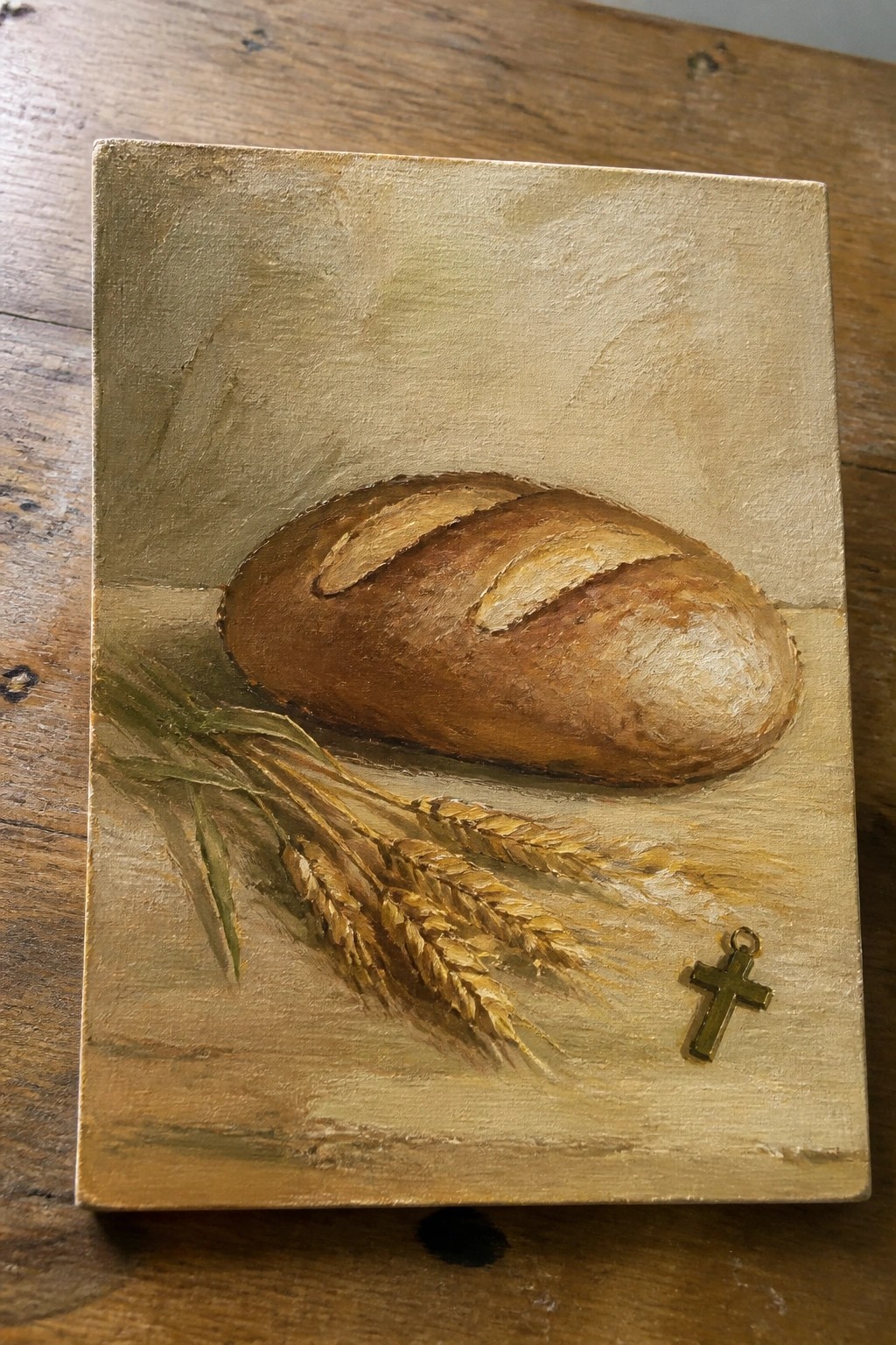 Still life painting of crusty bread loaf beside wheat stalks and small gold cross.
