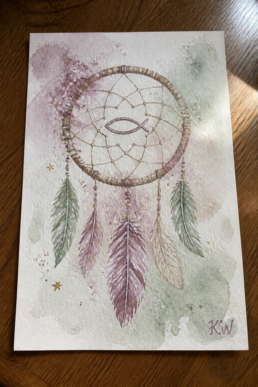 Watercolor dreamcatcher with fish symbol, dangling feathers, beads on purple-green background.