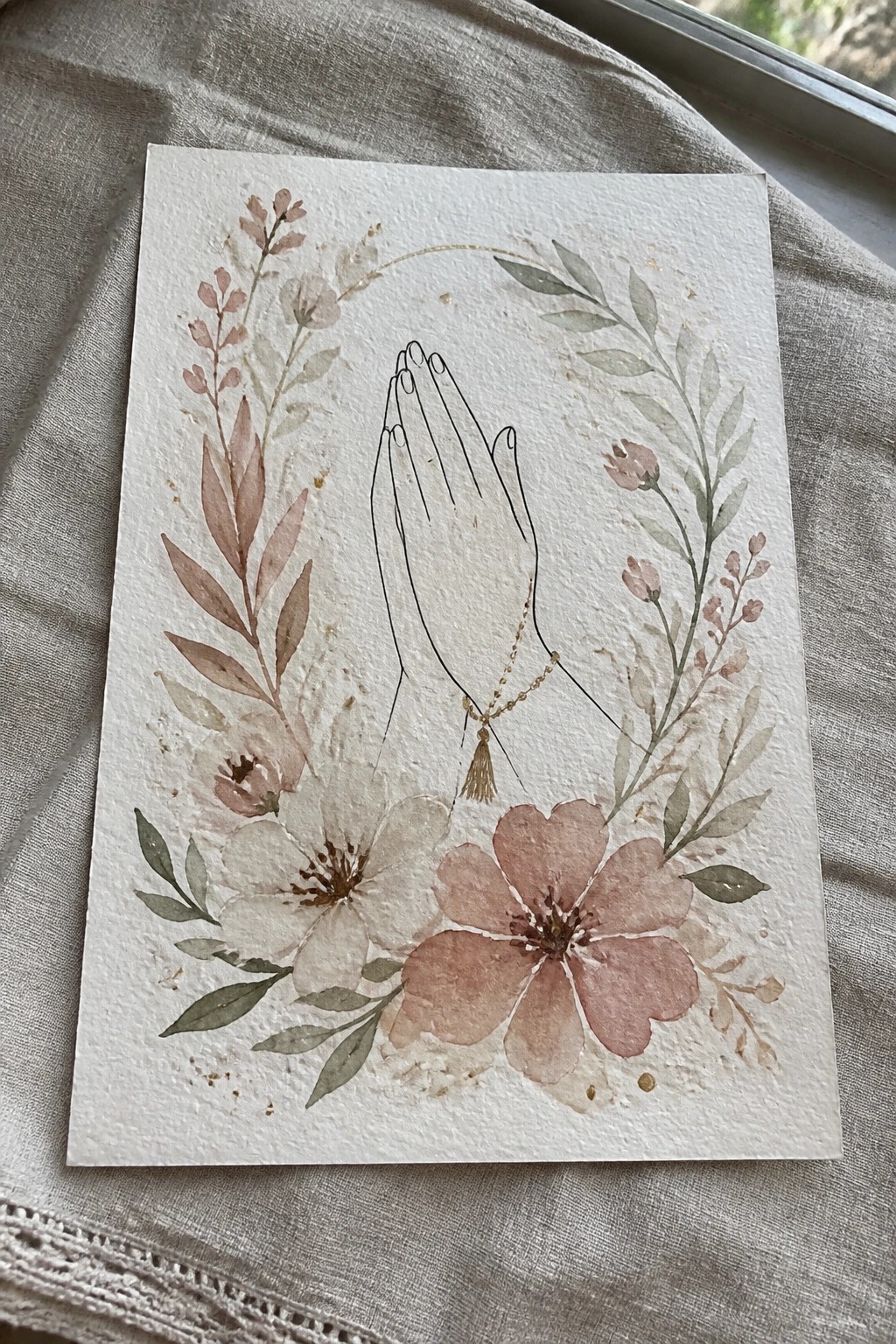 Watercolor illustration of praying hands with rosary necklace amid soft pink floral wreath
