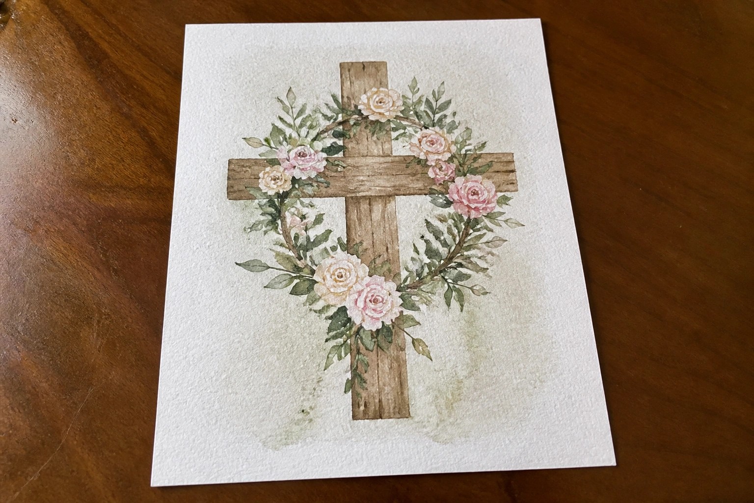 Watercolor painting of rustic wooden cross with pink and white roses and green leaves.