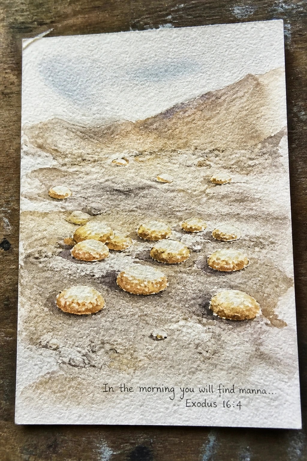 Watercolor of golden manna scattered on rocky desert ground with mountains and scripture text.