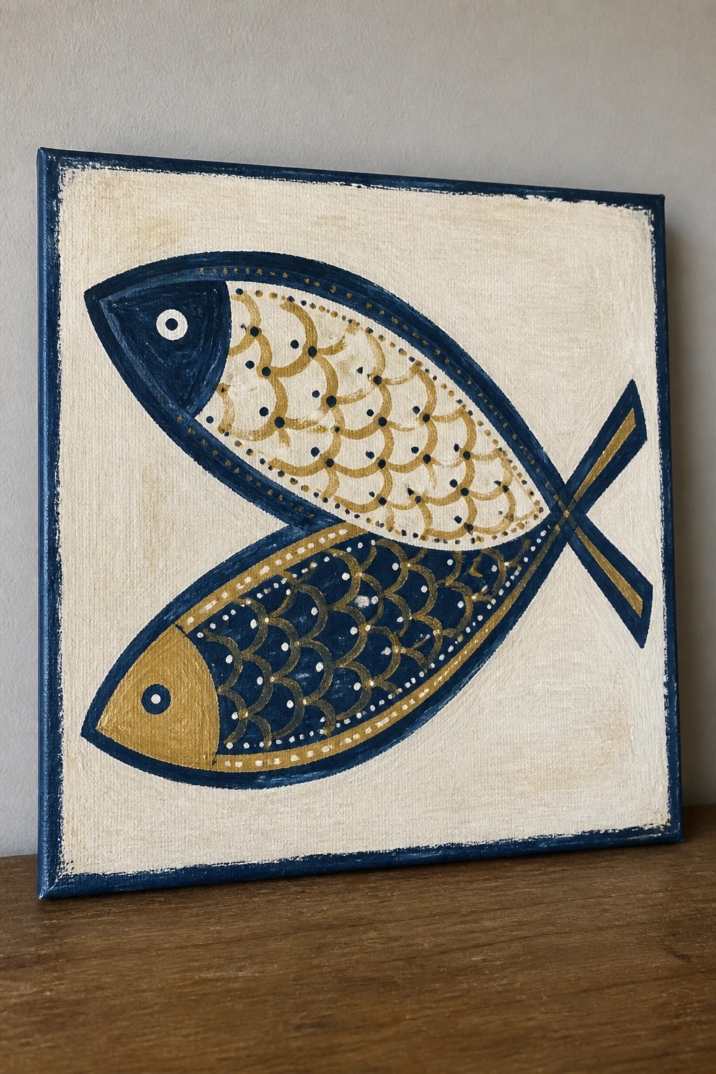A square canvas shows two stylized fish overlapping with tails crossed in an X, one with a dark head and the other with a gold head, both featuring scale patterns in gold and white on a cream background with navy borders.