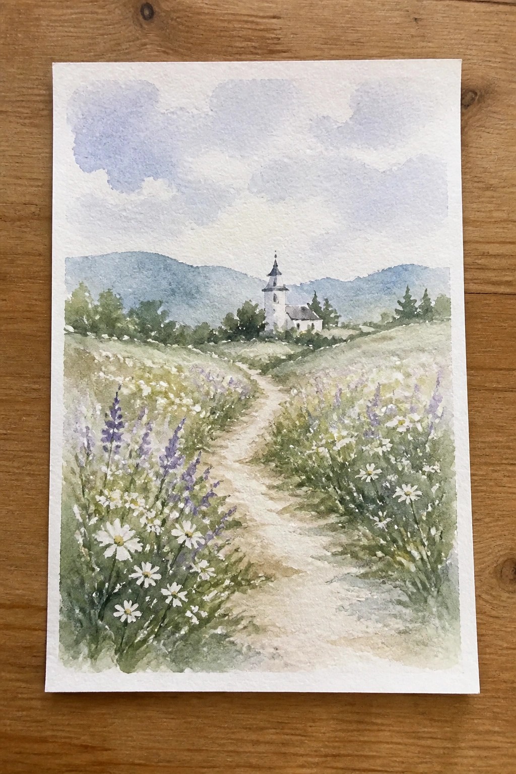 Watercolor of winding path through wildflowers leading to white church amid green hills.