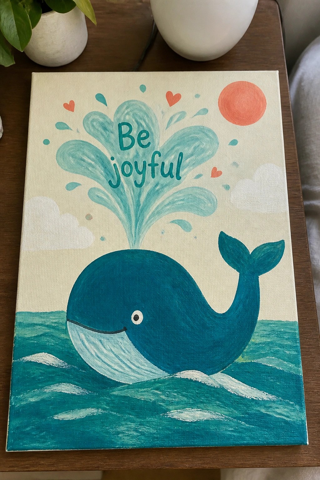 A canvas painting shows a blue whale in teal water with a water spout forming the words Be joyful, plus a red sun, hearts, and clouds.