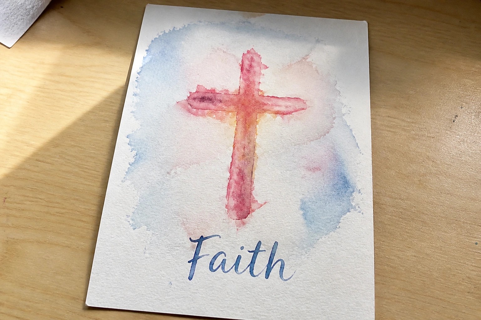 Watercolor cross in warm red-orange tones with blue-pink washes and cursive "Faith" below.