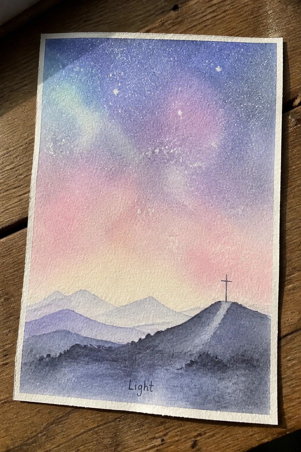Watercolor of starry purple-pink sky above dark mountains with cross and Light text