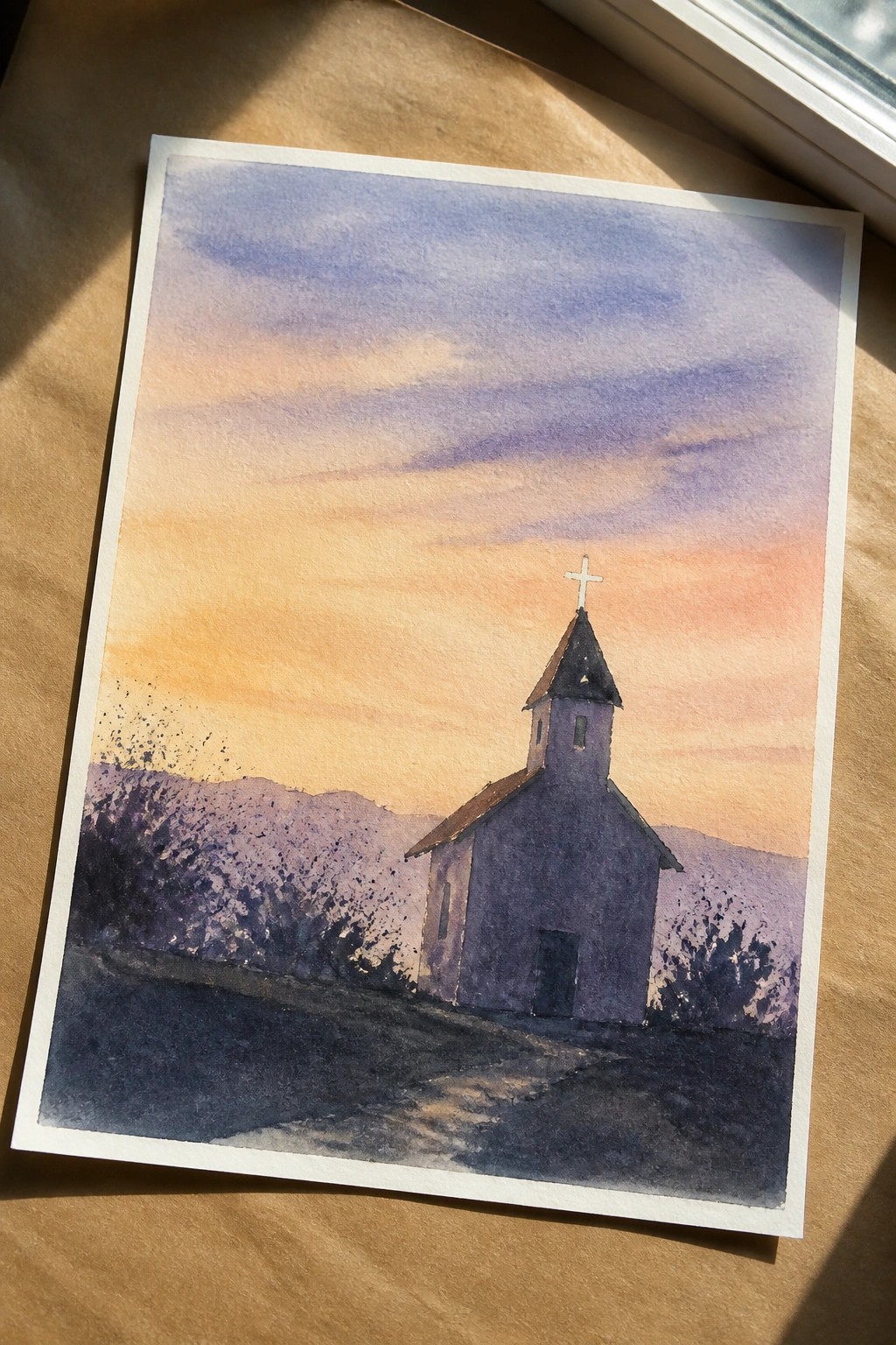 Watercolor painting of a small purple church with a cross-topped steeple on a hillside at sunset.