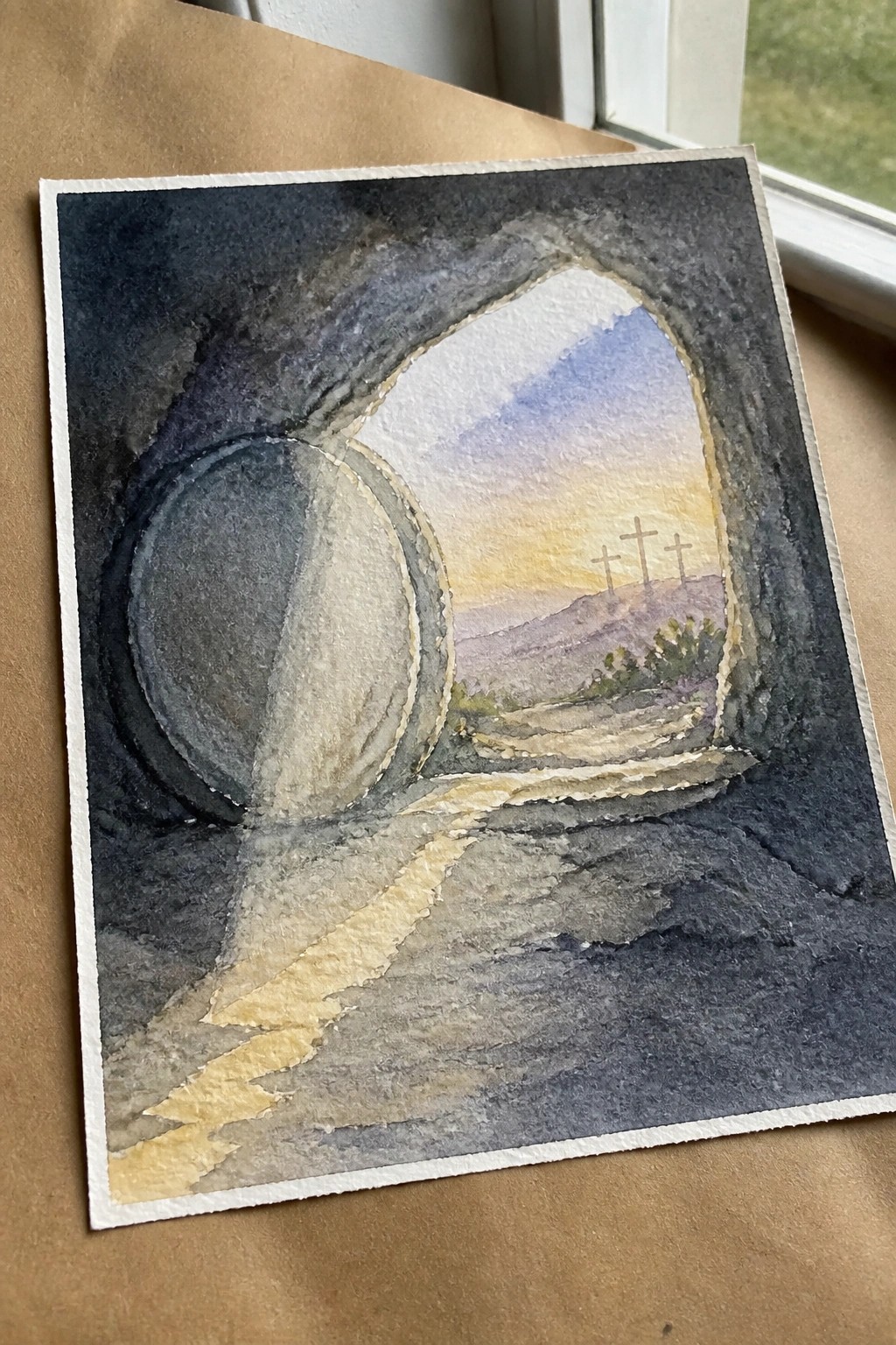 Watercolor of empty tomb with large stone rolled aside, three crosses on hill at sunrise.