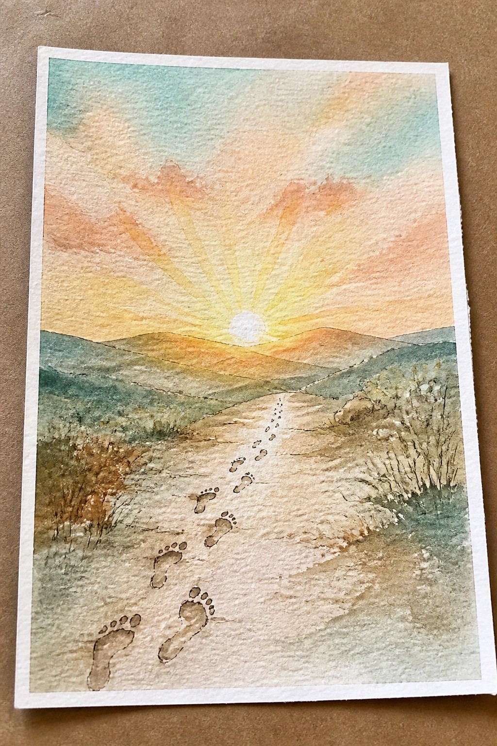 Watercolor painting of footprints on a winding path toward a sunset over mountains.