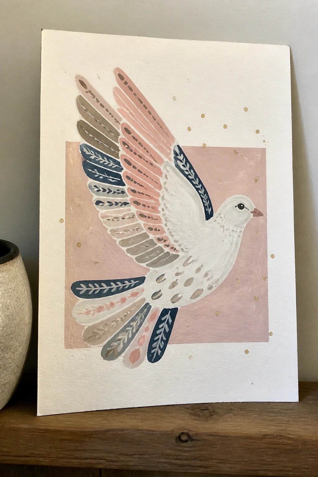 Watercolor painting of white dove with patterned pink and blue wings on pink square with gold dots.