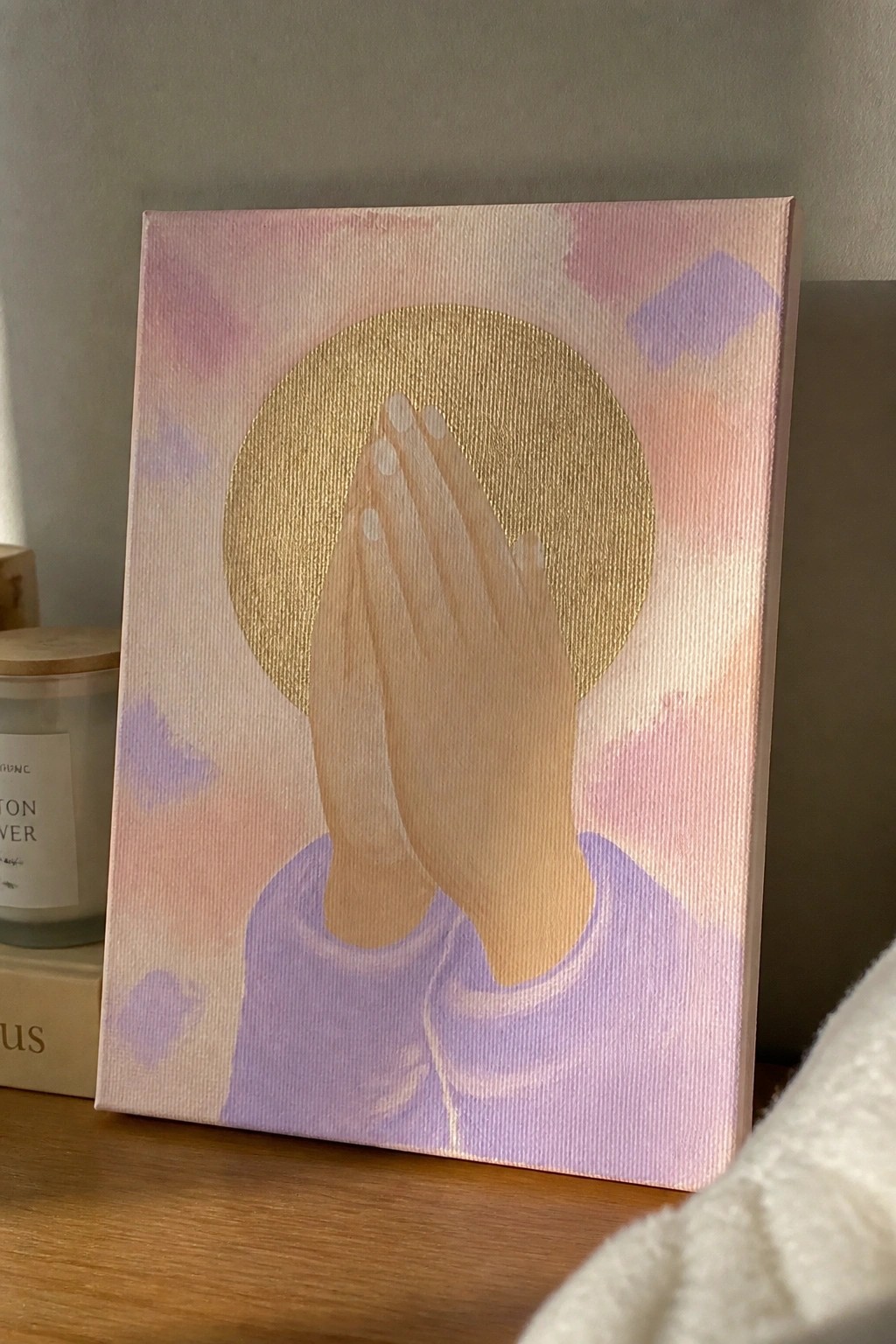 Painting of praying hands in purple top with gold halo on abstract pastel background.