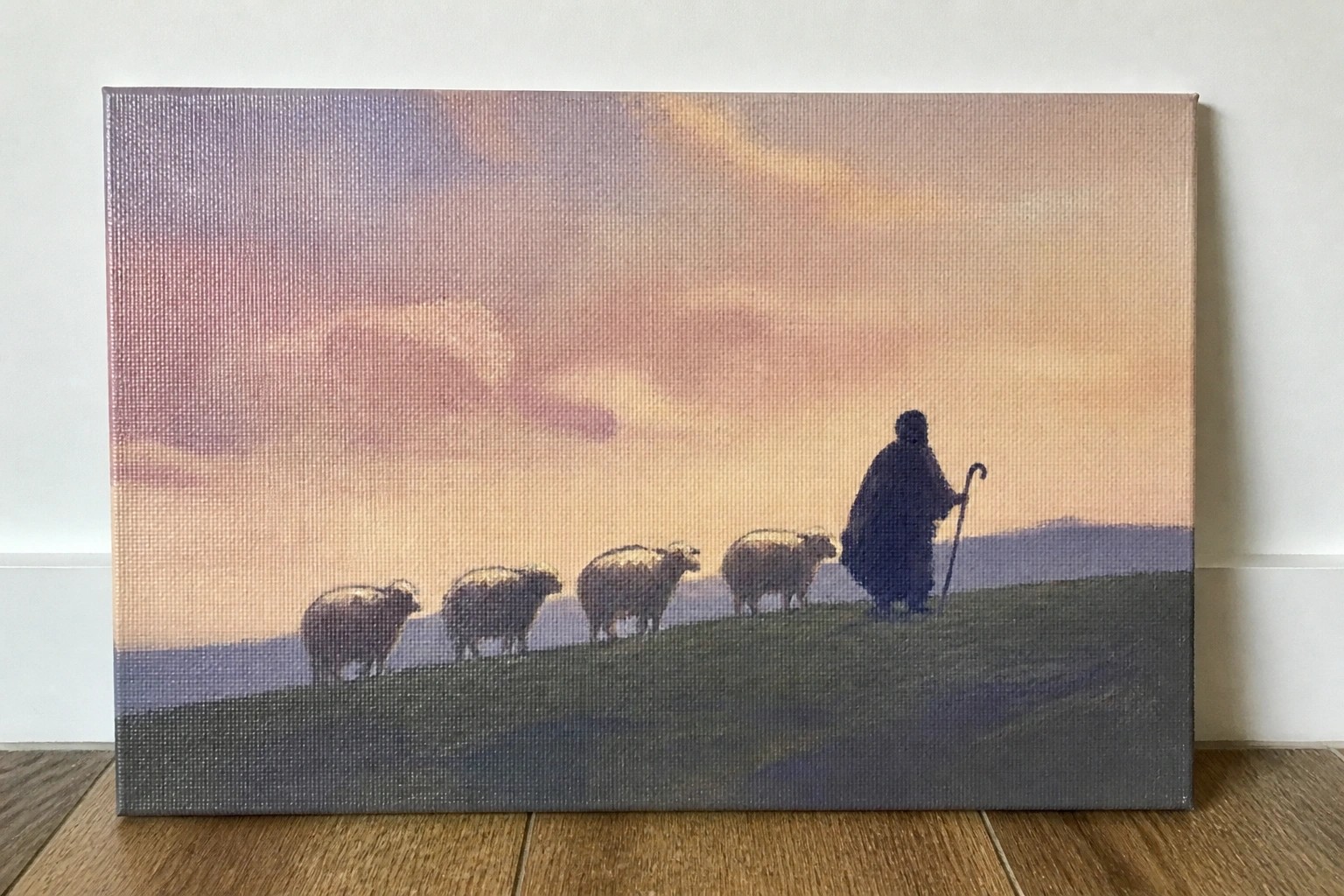 Silhouetted shepherd in dark cloak leads sheep across grassy hill under colorful sunset sky