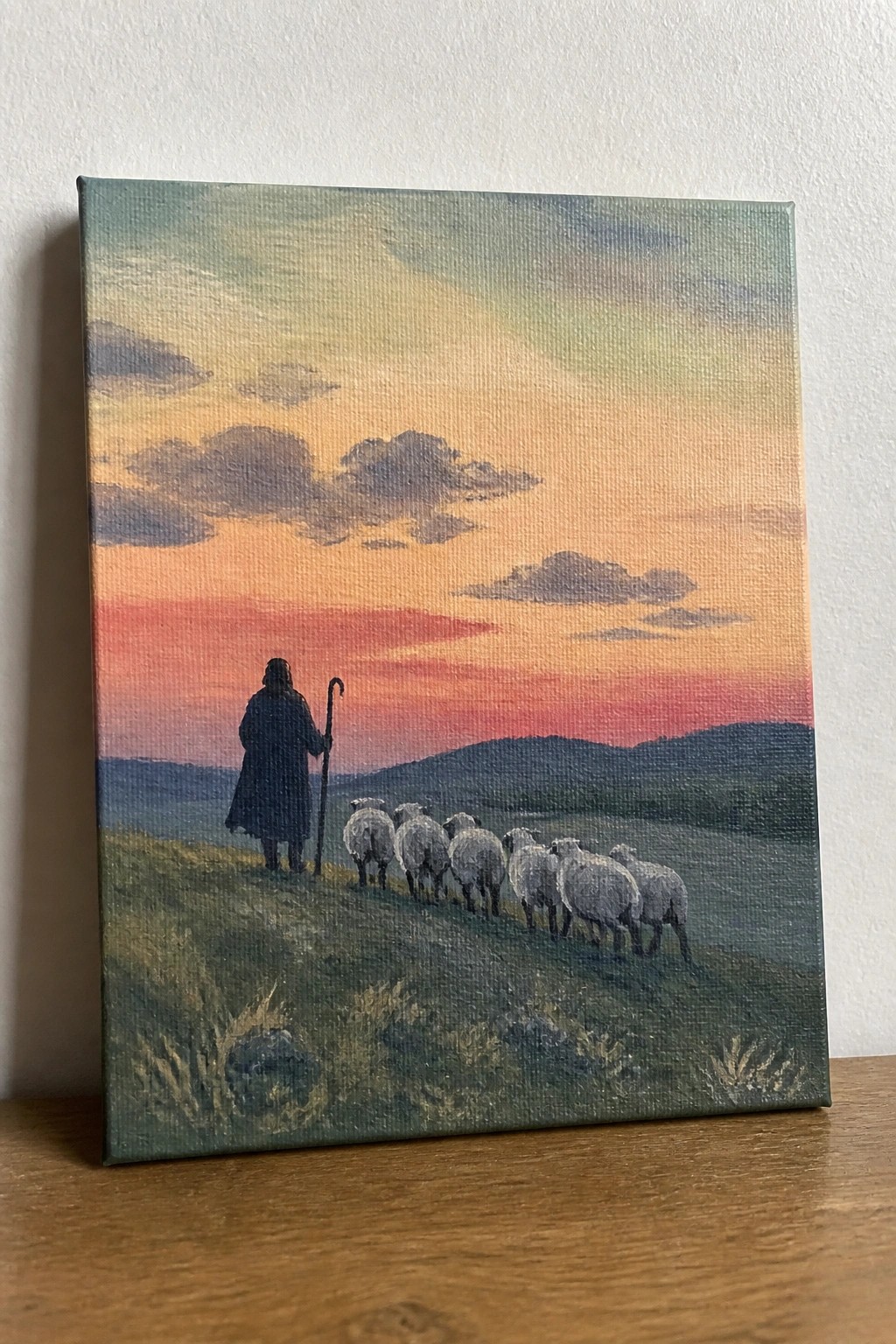 Silhouetted shepherd in dark cloak leads sheep across grassy hill under colorful sunset sky