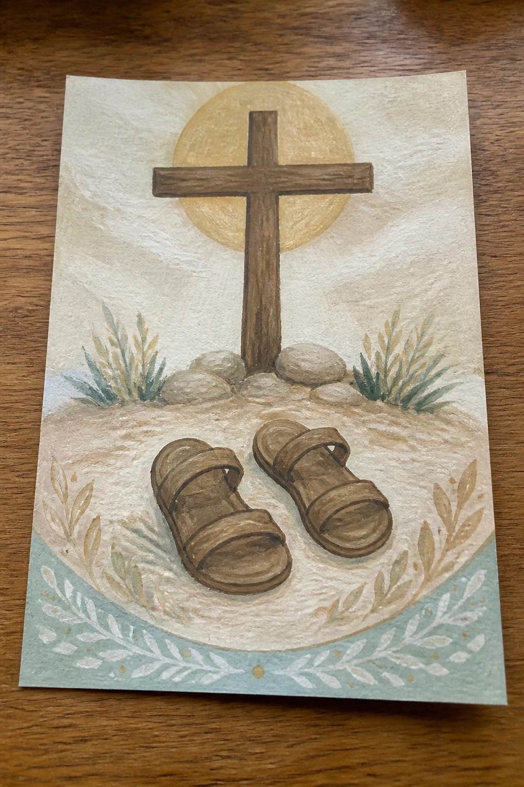Watercolor of wooden cross above sandals on sand, yellow sun, rocks, and plants.