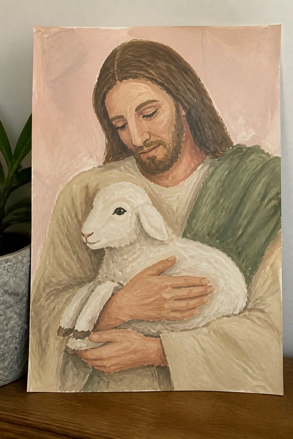 Jesus in green robe holds white lamb, long brown hair, pink background, painted style.