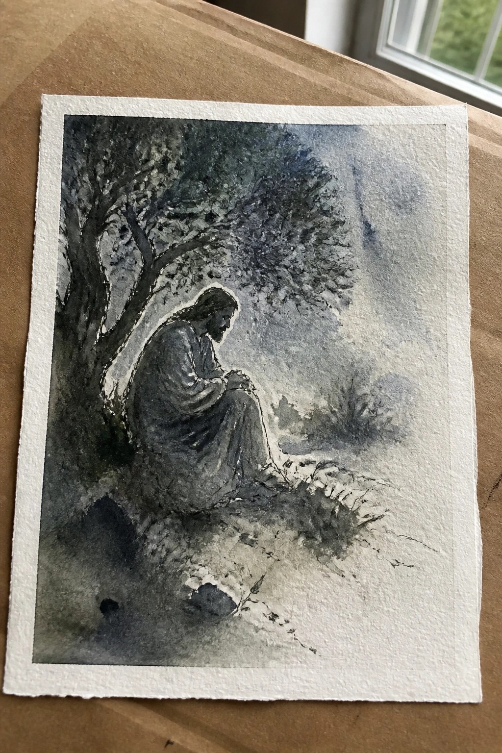 A watercolor painting of Jesus seated under a tree in a natural landscape, head bowed and hands clasped.