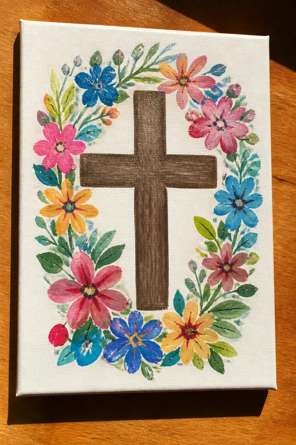 Brown cross centered on white canvas amid colorful floral wreath with green leaves.