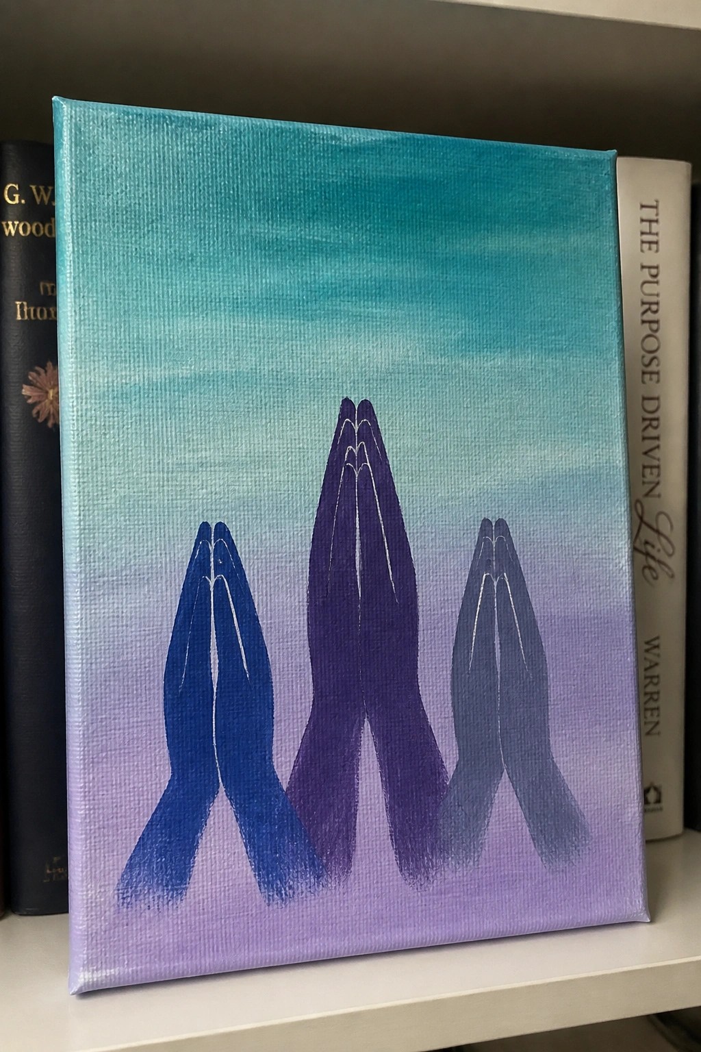 Canvas painting of three blue-purple praying hands on turquoise-purple gradient, between books.