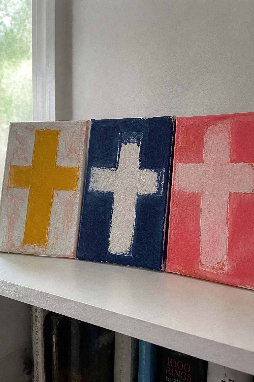 Three small canvases with yellow, white, and pink crosses on a white shelf.