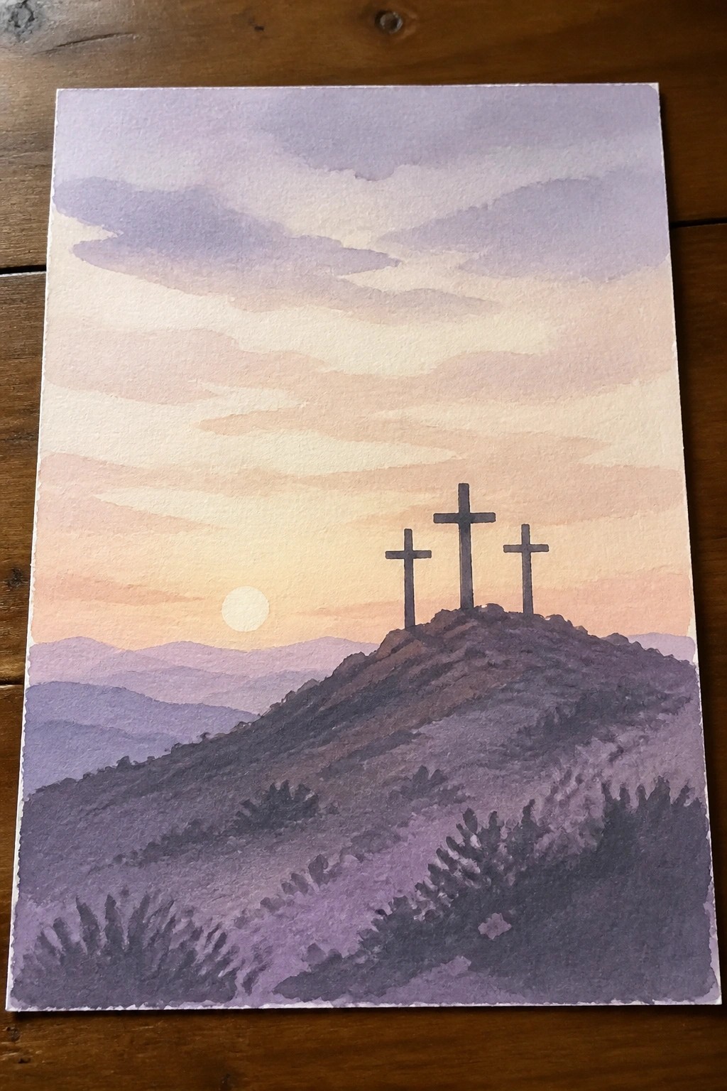 Three crosses on a hill under a purple-orange sunset sky with distant mountains.