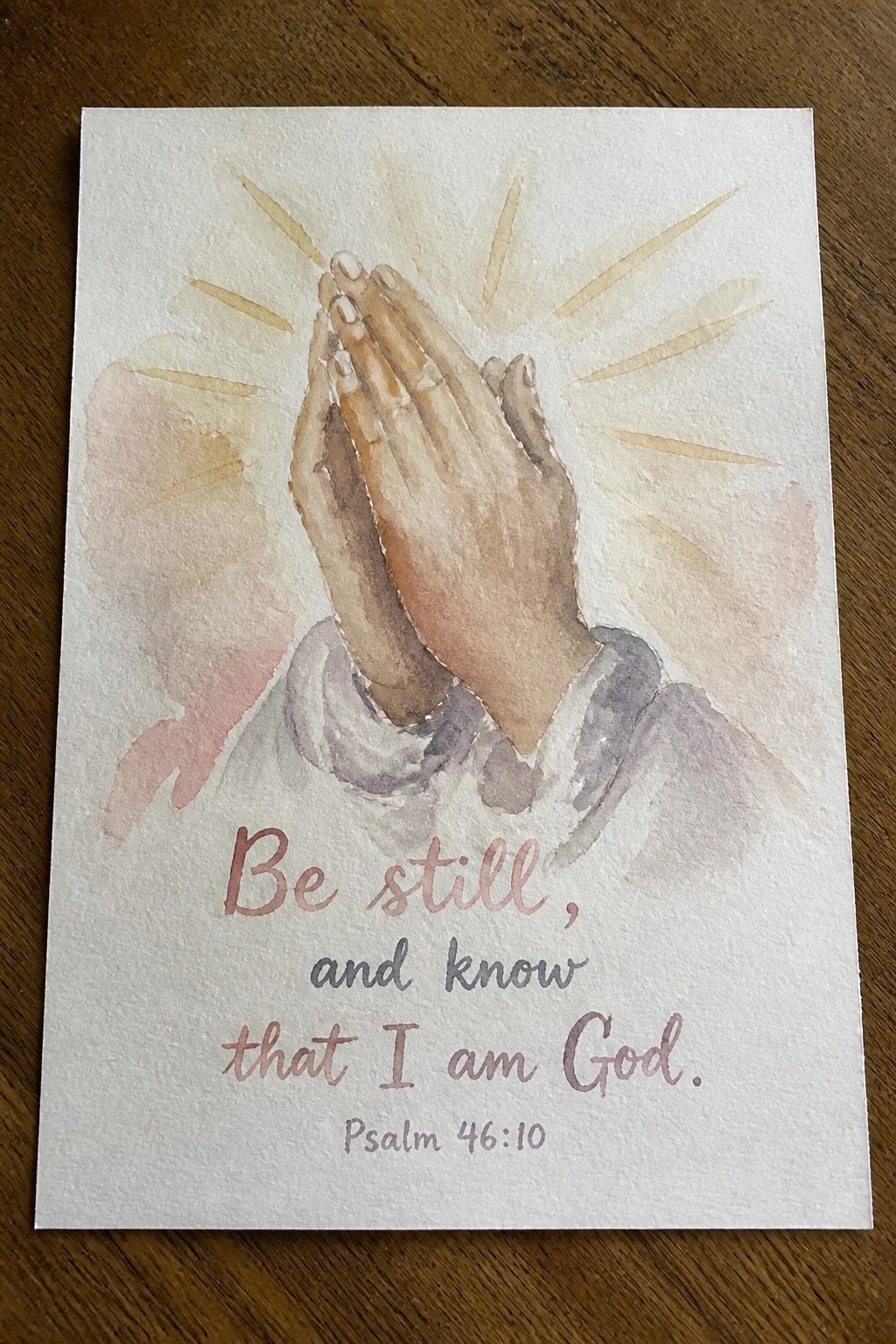 Watercolor of clasped praying hands with light rays, sleeves, and scripture text