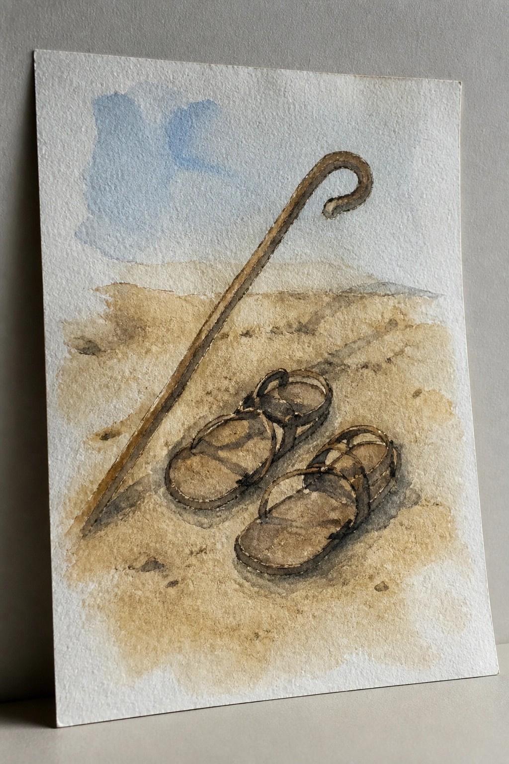 Watercolor of sandals and a shepherd's crook on sandy ground under blue sky.